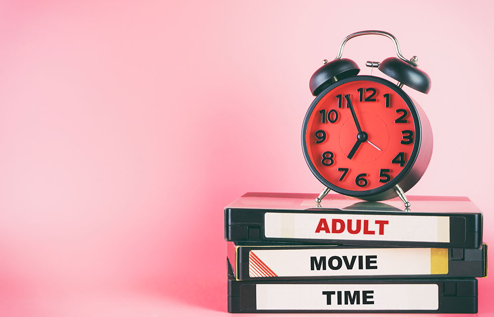 Wecker mit rotem Ziffernblatt auf 3 Videokassetten mit Beschriftung Adult Movie Time